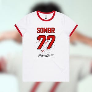 Pode incluir: Uma t-shirt branca de manga curta com acabamento vermelho na gola e nas mangas. A frente apresenta o texto SOMBR acima do número 77 em vermelho com contorno preto e um efeito de salpicos, seguido por uma assinatura gráfica preta.