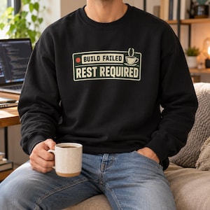 May include: A black long-sleeve crewneck sweatshirt featuring a rectangular graphic with a red dot, a coffee cup icon, and the text "BUILD FAILED" above "REST REQUIRED" in a light cream colour, printed on the centre of the chest.