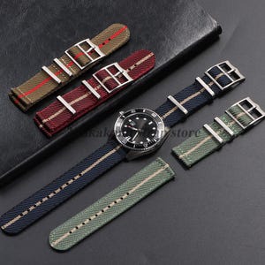 May include: A stainless steel wristwatch with a black dial and a navy blue fabric strap featuring a central beige stripe, accompanied by three additional fabric watch straps in olive green, burgundy, and khaki, each with silver-toned metal buckles.