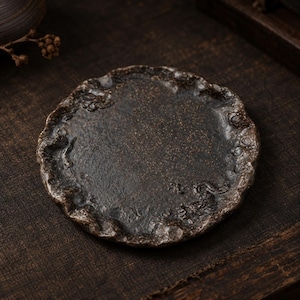 Peut inclure: Une soucoupe ronde en céramique brun foncé avec un bord texturé et irrégulier. La surface présente une finition mouchetée et des motifs de dragon en relief intégrés dans la bordure surélevée et rugueuse, offrant un aspect terreux et patiné.