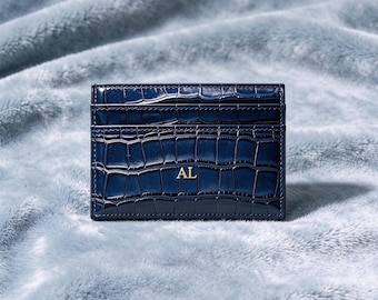 May include: A rectangular navy blue card holder with a glossy crocodile skin texture. The front features two horizontal card slots and the gold-colored embossed letters AL centered in the middle of the lower section of the accessory.