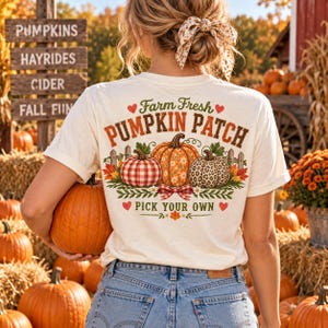 Puede incluir: Una camiseta color crema con un gráfico en la espalda que incluye el texto Farm Fresh PUMPKIN PATCH PICK YOUR OWN. El diseño muestra tres calabazas con diferentes patrones, corazones rojos y hojas verdes, combinada con pantalones vaqueros azules.