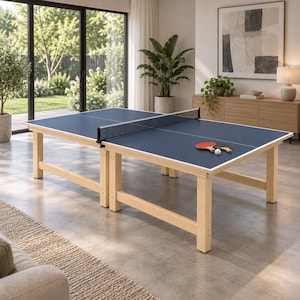 Puede incluir: Una mesa de ping pong rectangular con superficie de juego azul oscuro, líneas blancas en los bordes y una red negra en el centro. La mesa cuenta con patas y estructura de madera clara, con una pala roja y negra y una pelota blanca encima.