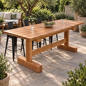May include: A rectangular wooden dining table with a natural finish and a trestle base, accompanied by four black metal bar stools. The table surface features horizontal planks and holds a small gray bowl with greenery and two clear glasses.