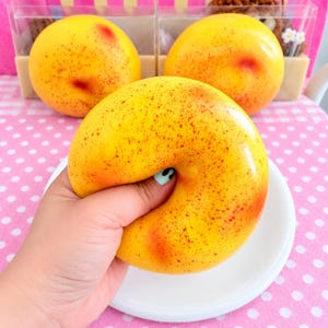 May include: Three round, yellow, peach-shaped squishy toys with red speckles and a smooth, glossy finish. One toy is held in the foreground, while two others sit on a white plate in the background, all featuring a soft, squeezable texture.