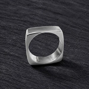 Könnte beinhalten: Ein silberfarbener Ring mit einem markanten, quadratischen Außenband und einer glatten, polierten Oberfläche. Der Innenumfang des Rings ist kreisförmig, was einen Kontrast zwischen dem kantigen Äußeren und dem abgerundeten Innendesign erzeugt.
