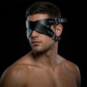 Puede incluir: Un antifaz de cuero negro con dos correas cruzadas que cubren los ojos. El diseño incluye hebillas de metal plateado, remaches y una anilla circular en el lateral, asegurado con correas ajustables alrededor de la cabeza.