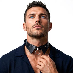 Op de afbeelding: Een zwarte choker van lederachtig materiaal met een puntig, hemdkraag-achtig ontwerp, voorzien van zilverkleurige metalen studs langs de band en een centrale zilverkleurige metalen ring aan de voorzijde van de hals.