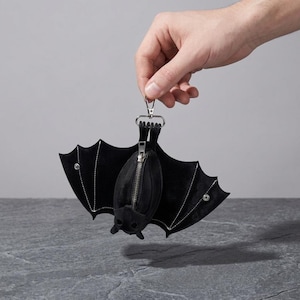May include: A black bat-shaped coin purse featuring a central silver-toned zip closure, white stitched detailing along the wings, and a metal lobster clasp attachment at the top. The wings are secured with small silver-toned snap buttons.