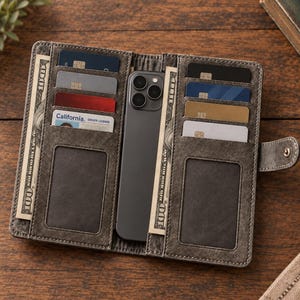 Puede incluir: Una funda tipo cartera gris con textura desgastada que sostiene un teléfono inteligente en el centro, con múltiples ranuras para tarjetas a ambos lados. La cartera contiene varias tarjetas bancarias, una licencia de conducir de California y dos billetes de 100 dólares.