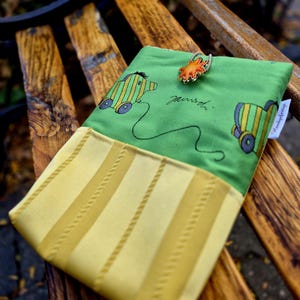 Könnte beinhalten: Eine grüne Stofftasche mit einem gelb gestreiften unteren Bereich und einem Aufdruck eines kleinen Spielzeugs auf Rädern. Die Tasche wird mit einer elastischen Schlaufe und einem sternförmigen Knopf verschlossen. Ein weißes Etikett zeigt Handgefertigt.