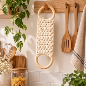 Op de afbeelding: Een crèmekleurige macraméband met een gestructureerd knopenpatroon, aan beide uiteinden verbonden met gladde, ronde, lichtgekleurde houten ringen. De band hangt aan een houten wandhaak en toont een functioneel ontwerp met natuurlijk koord.