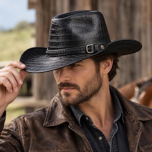 Puede incluir: Un sombrero de vaquero negro con un patrón texturizado de piel de cocodrilo en la copa y el ala. El sombrero incluye una banda trenzada decorativa, una hebilla de metal plateado y un pequeño detalle de tachuela metálica circular en la banda.