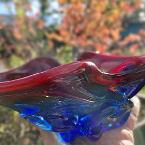 Può includere: Una ciotola in vetro soffiato a mano caratterizzata da un vivace gradiente di colore che sfuma da un blu profondo e saturo alla base a un rosso ricco e traslucido lungo il bordo ondulato e organico, con una finitura liscia e lucida.