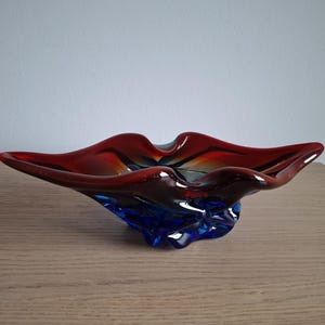 Puede incluir: Un cuenco decorativo de cristal con un borde ancho y ondulado de color rojo intenso que se transforma en una base de color azul vibrante. El cuenco tiene una superficie lisa y pulida con curvas orgánicas fluidas y una calidad translúcida.