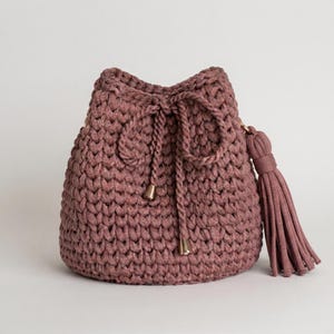 May include: A dusty rose bucket bag crafted from thick, chunky crochet yarn. The bag features a drawstring closure with two braided cords ending in metallic gold-toned caps, and a large, matching tassel attached to the side of the bag.