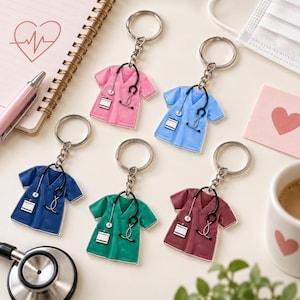 May include: Five acrylic keychains shaped like scrub tops in pink, light blue, dark blue, green, and maroon. Each keychain features a printed black stethoscope and a white rectangular ID badge attached to a silver metal split ring and chain.