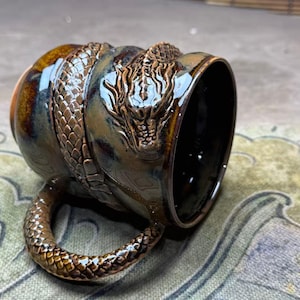 May include: A ceramic mug with a glossy brown and black glaze featuring a three-dimensional sculpted dragon wrapped around the exterior. The dragon's tail forms the handle, and its detailed head is prominently displayed on the side.