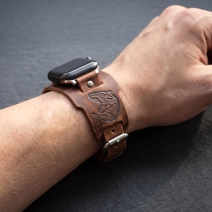 Può includere: Un cinturino per orologio in pelle marrone con un design a polsino largo e una grafica a testa di lupo in rilievo. Il cinturino è fissato a una cassa per smartwatch rettangolare nera e include una fibbia in metallo argentato e un rivetto.