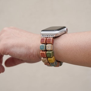 May include: A silver smartwatch featuring a double-strand watch band made of small, square, ceramic-style beads in various earthy tones including terracotta, teal, mustard yellow, and speckled cream, connected by small silver-toned metal spacers.