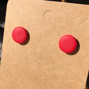 May include: A pair of bright red, round stud earrings on a brown earring card. The card is printed with the words "Handmade with love". The earrings are a simple, minimalist design, suitable for everyday wear.