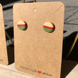 May include: A pair of handmade stud earrings on a brown card. The earrings are round with a beige top, a red stripe, and a green bottom. The card reads "Handmade with love."
