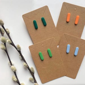May include: Four pairs of stud earrings in green, orange, and blue, displayed on brown cardboard earring cards. The earrings are small, rectangular shapes. A sprig of pussy willow branches is in the background.