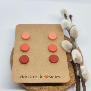 May include: A set of six round stud earrings in shades of peach, coral, and red-brown, displayed on a brown card. The card reads "Handmade with love." The earrings are arranged in pairs, showcasing a gradient of colors.