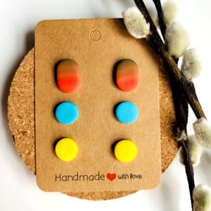 May include: A display of six colourful stud earrings on a brown card. The earrings are arranged in three pairs: orange-red gradient ovals, bright blue circles, and yellow circles. The card reads "Handmade with love."