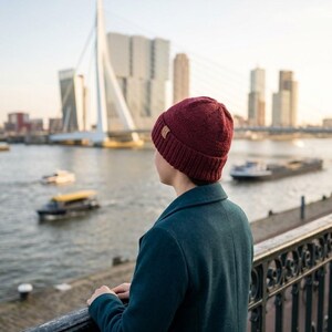 Op de afbeelding: Een diepbordeauxrode gebreide muts met een geribbelde boord en een klein vierkant bruin lederachtig label aan de zijkant. De muts heeft een textuurrijke gebreide constructie en wordt op het hoofd gedragen, waarbij het breipatroon zichtbaar is.