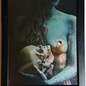Könnte beinhalten: Ein gerahmter Kunstdruck, der eine Figur mit blau-türkisen Hauttönen zeigt, die einen menschlichen Schädel hält. Die Figur weist metallische Goldakzente auf der Brust und am Schädel auf, mit langem dunklem Haar vor einem dunklen Hintergrund.