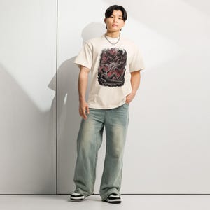 May include: A cream-colored short-sleeve t-shirt featuring a dark, detailed graphic print on the chest. The illustration depicts a winged, demonic figure hovering above a reclining person reaching upward, set against a backdrop of swirling red smoke.
