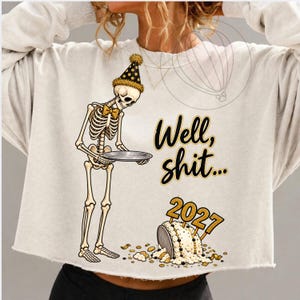 May include: A cream-colored cropped sweatshirt featuring a graphic of a skeleton wearing a gold and black party hat and bow tie, holding a silver platter. The text reads "Well, shit..." above a fallen cake with "2027" cake toppers.