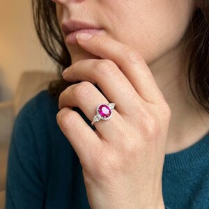 Anillo de oro blanco de 18 quilates con un rubí central, rodeado por un halo de pequeños diamantes blancos y dos diamantes en forma de pera a los lados.