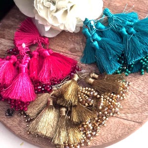 Può includere: Tre set di nappe decorative in rosa brillante, verde acqua e oro metallizzato. Ogni gruppo di nappe presenta molteplici fili di seta legati in cima, impreziositi da piccole perline abbinate che pendono dalla parte inferiore dei fili.