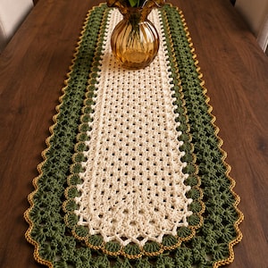 May include: A rectangular crochet table runner featuring a cream-colored center with a grid pattern, surrounded by a wide border of dark green yarn. The edges are finished with a scalloped design trimmed in a thin line of golden yellow yarn.