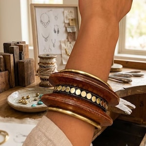 Puede incluir: Un conjunto de pulseras formado por dos brazaletes de metal dorado liso que flanquean un brazalete ancho de madera marrón oscuro. El brazalete de madera presenta una banda central negra incrustada con una fila de pequeños tachones metálicos dorados.