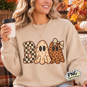 May include: A beige crewneck sweatshirt featuring the word BOO printed in large letters. The B is a black and beige checkerboard pattern, the O is a ghost with orange stars, and the second O is a ghost with orange flowers and a polka dot bow.