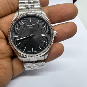 May include: A silver-tone Tissot 1853 wristwatch featuring a black sunray dial with silver indices, a date window at the three o'clock position, a fluted bezel, and a multi-link metal bracelet partially wrapped in protective plastic film.