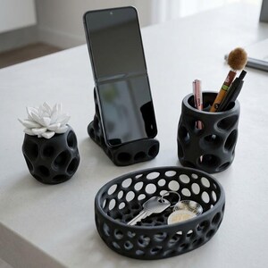 Può includere: Un set di accessori da scrivania neri con una texture porosa e organica, che include un piccolo vaso con una succulenta bianca, un supporto per smartphone con un telefono nero, un portapenne cilindrico e un vassoio ovale con una chiave metallica.
