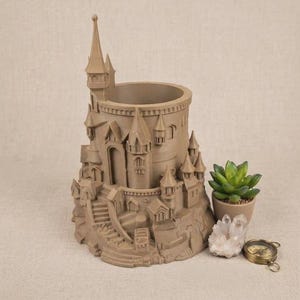 May include: A light brown, cylindrical castle-shaped planter featuring multiple turrets, a central tower, and a staircase. Beside the planter sits a small green succulent in a pot, a cluster of clear quartz crystals, and a small brass compass.