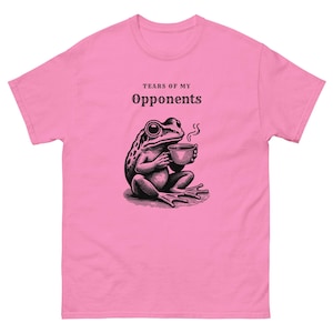 Könnte beinhalten: Ein hellrosa T-Shirt mit kurzen Ärmeln und einer schwarzen grafischen Illustration eines Frosches, der eine dampfende Tasse hält. Über dem Frosch steht der Text "TEARS OF MY OPPONENTS" in einer fetten, dunklen Schriftart.