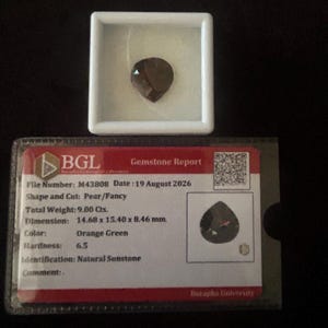 Può includere: Una pietra di sole naturale a forma di pera di colore arancione-verde, presentata in una custodia quadrata bianca accanto a un rapporto gemmologico BGL. Il rapporto indica dimensioni di 14,68 x 15,40 x 8,46 mm, un peso di 9,00 carati e una durezza di 6,5.