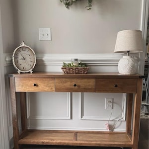 May include: A rectangular wooden console table with three drawers and a lower shelf. On top sits an oval-shaped white analog clock with black numerals and a small rectangular woven basket containing greenery, alongside a white textured table lamp.