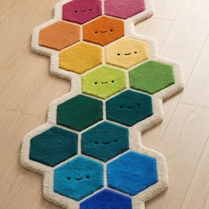 May include: A runner rug composed of interconnected hexagonal segments in a rainbow gradient of colours. Each hexagon features a simple embroidered face with two dots for eyes and a curved line for a mouth, all outlined in a cream-coloured border.
