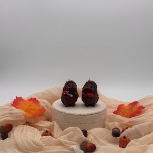 May include: Two brown hedgehog-shaped candles standing on a circular beige platform. One hedgehog holds a small red mushroom, while the other holds a red heart. The scene is decorated with orange and yellow fabric leaves, small acorns, and tiny pumpkins.