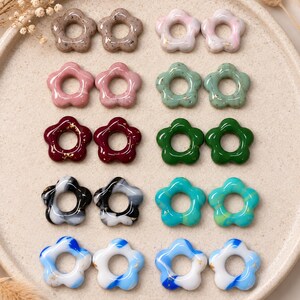 May include: A collection of ten pairs of small, flower-shaped beads with a glossy finish, arranged in five rows of two pairs each. The beads feature various colours including speckled beige, pale pink, sage green, deep red, dark green, black, turquoise, and blue.