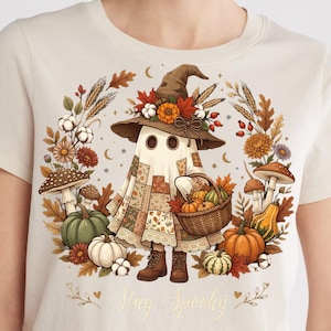Pode incluir: Uma t-shirt cor de creme com um gráfico de um fantasma com uma capa de retalhos, um chapéu de bruxa adornado com flores e botas castanhas. O fantasma carrega um cesto de abóboras, rodeado por folhas de outono, cogumelos e o texto Stay Spooky.