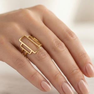 Könnte beinhalten: Ein goldfarbener Ring mit einem offenen, geometrischen Design, das aus drei übereinanderliegenden rechteckigen Rahmen besteht. Der Ring hat eine polierte Oberfläche und wird am Ringfinger getragen, wobei die hohle Struktur hervorsticht.