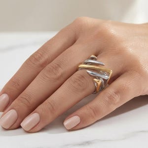 May include: A two-tone ring featuring a combination of polished silver-colored and gold-colored metal bands. The design consists of multiple overlapping, curved, and angular segments that create a layered, dimensional appearance on the finger.